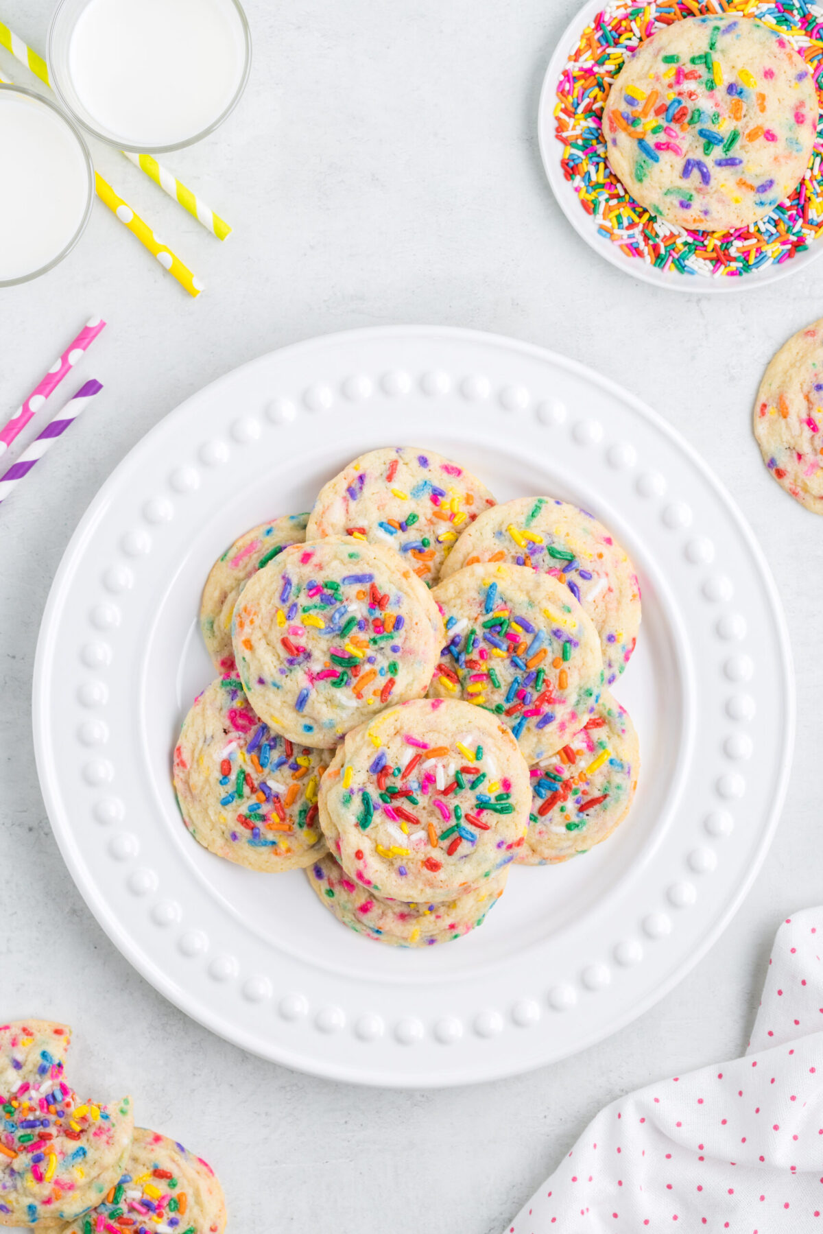 cookies spread out on a white plate in the centre, cookies top right with mor sprinkles circling the cooking, 2 glasses of milks top lip (top down view) with yellow, red and purple striped straws, more cookies bottom left and white linen bottom right
