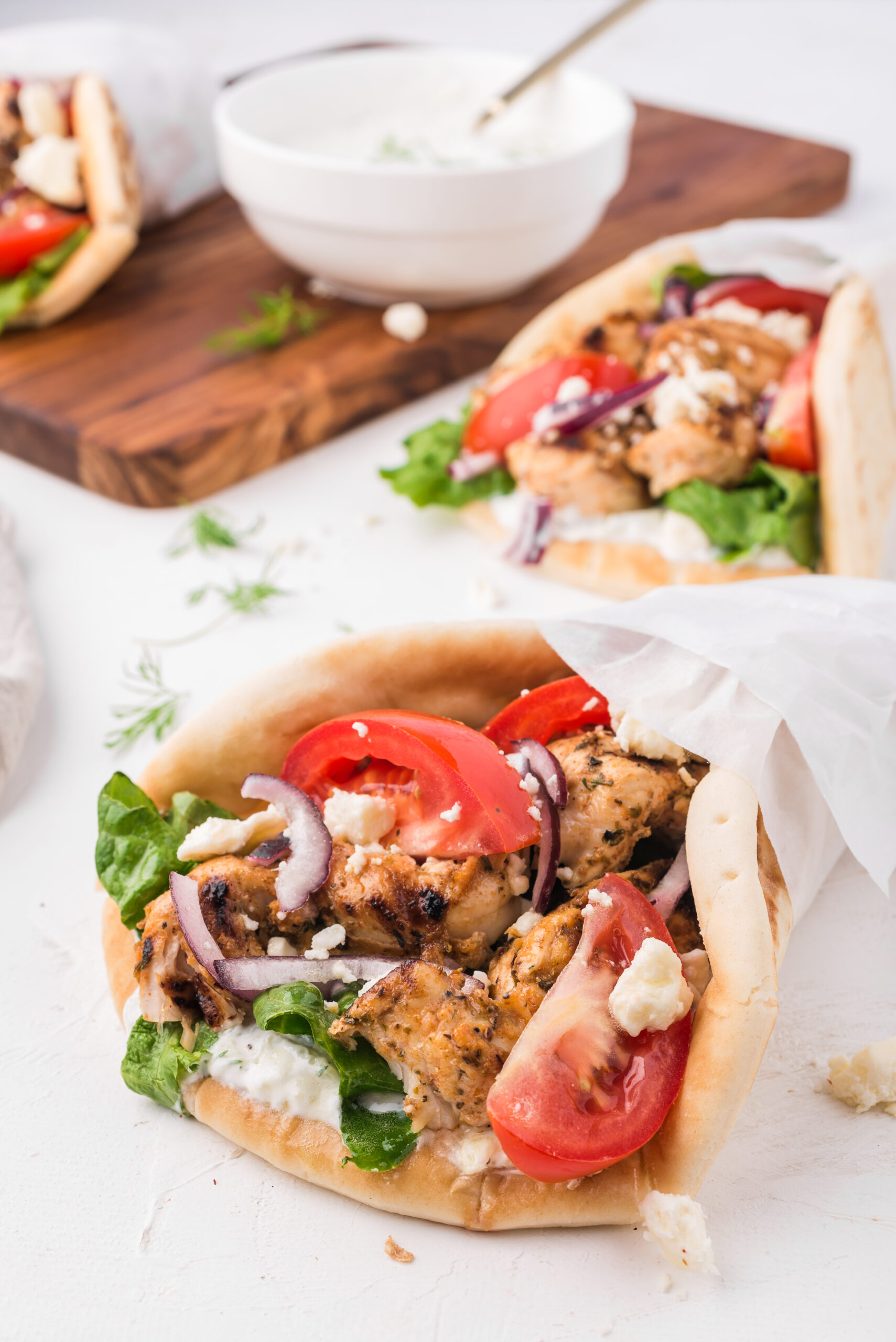 Grilled Chicken Gyros, one close up wrapped in parchment paper showing chicken, tomato, purple onions, feta cheese. second gyro to the back right, back left wooden chopping board with a white bowl with tzatziki and to the left a third gyro