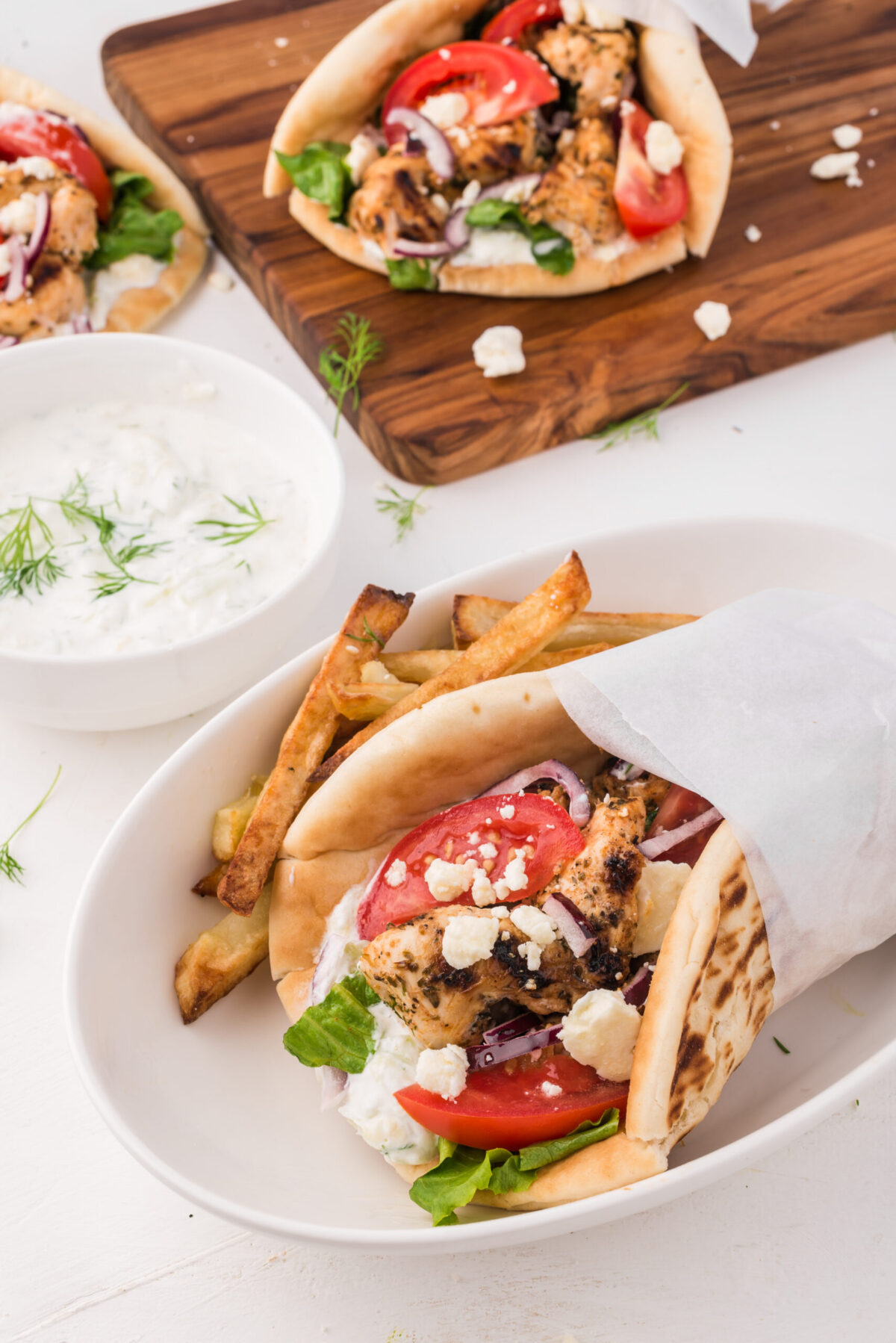 gyros assembled, wrapped in parchment paper (white) on a white deep plate with fries on the left of the gyros. White bowl with tzatziki on the left of the plate with chopped dill on top. Top middle right wood cutting board with a gyro