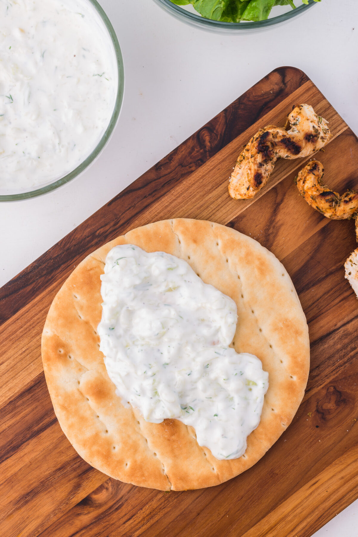 pita bread on a woo cutting board with tzatziki spread on it, grilled chicken pieces top right, tzatziki in a glass bowl top left
