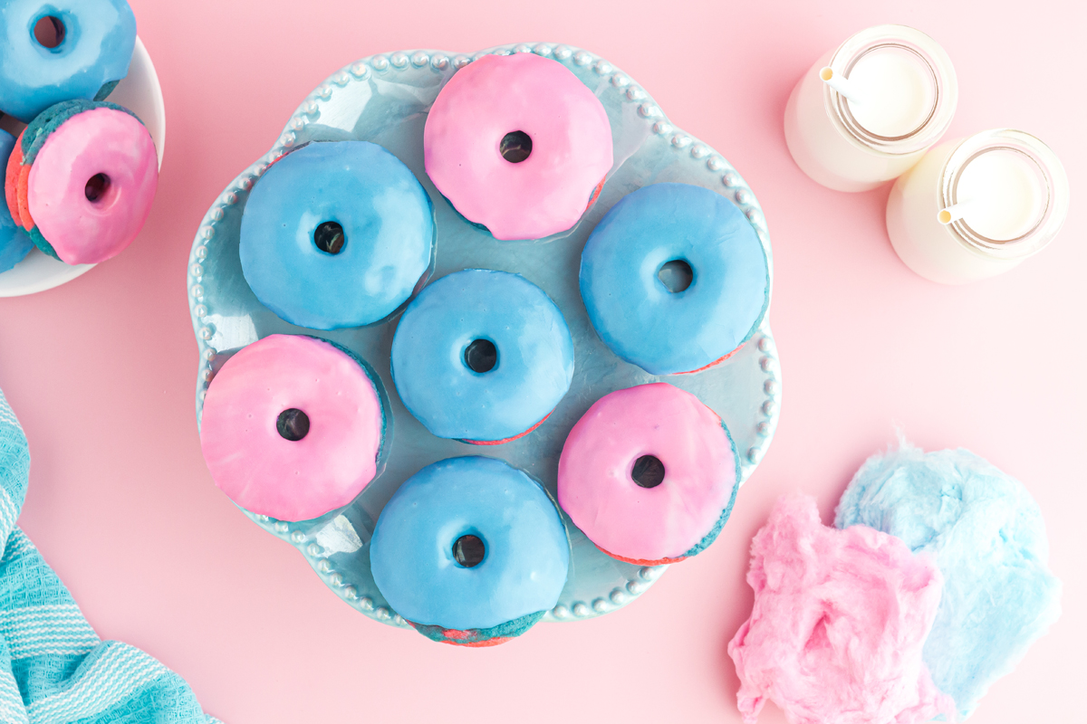landscape photo 4 blue & 3 pink donuts on a blue plate with donuts in the top left, milk jugs on the top right and bottom right cotton candy peeking out all on a pink background.