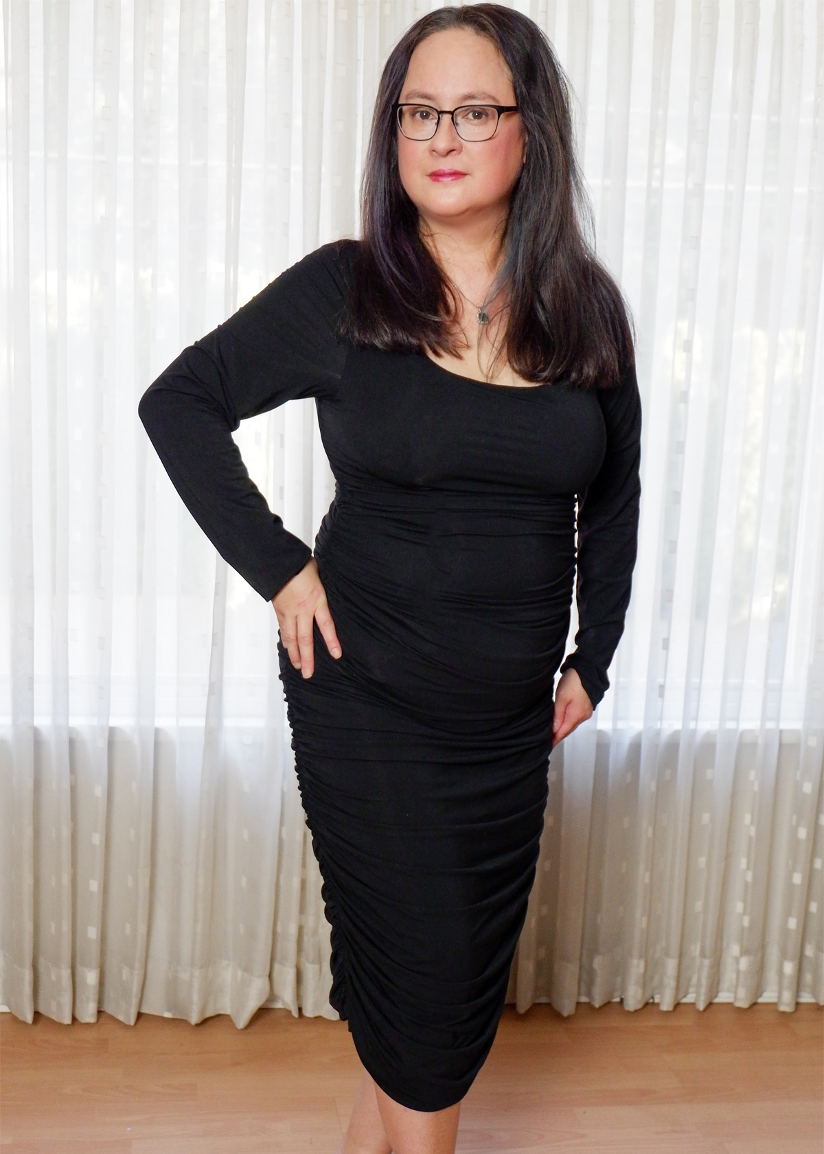 powered by Mom wearing black framed glasses, black ruched shaping dress. Right hand on hip, left arm straight down.