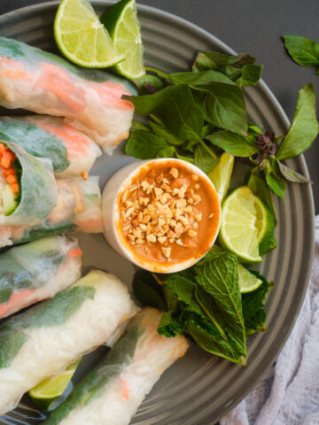 Vietnamese Peanut Sauce with crushed peanuts on top in a single dipping bowl surrounded with Thai Basil, min, lime wedges and lemongrass chicken salad rolls